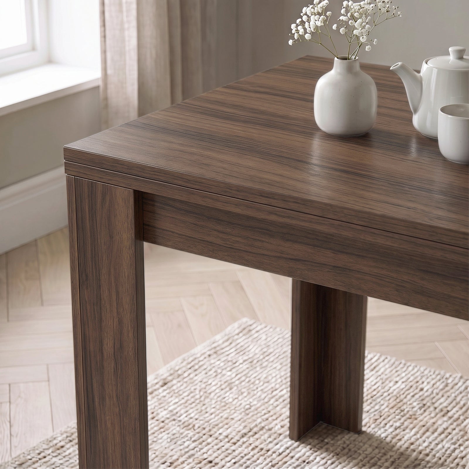 [Walnut] Close-up of the folded wood-grain square tabletop above apron and wide legs, with white flower vase, teapot and cup