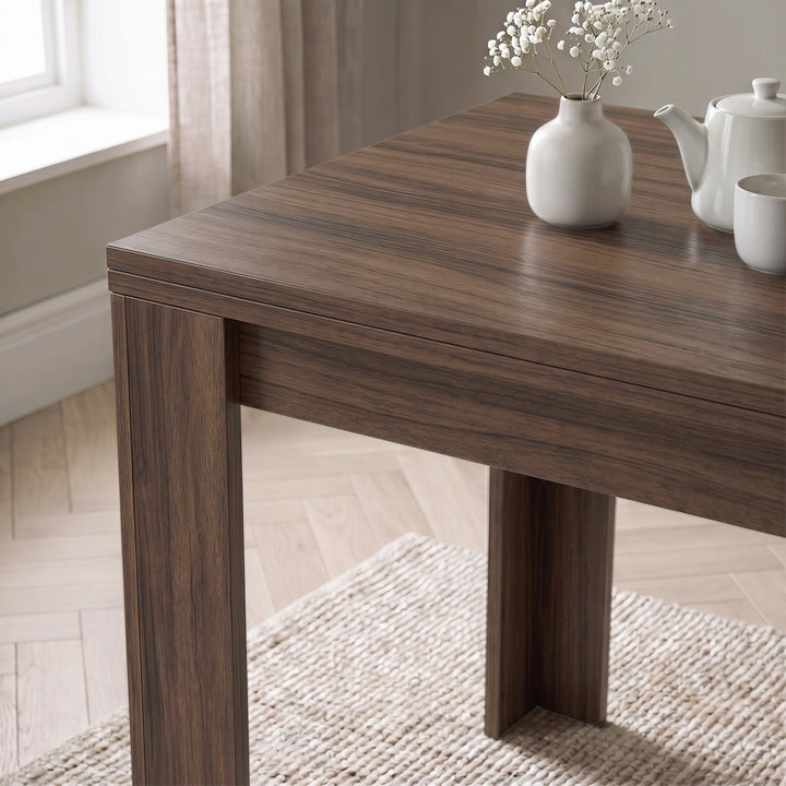 [Walnut] Close-up of the folded wood-grain square tabletop above apron and wide legs, with white flower vase, teapot and cup