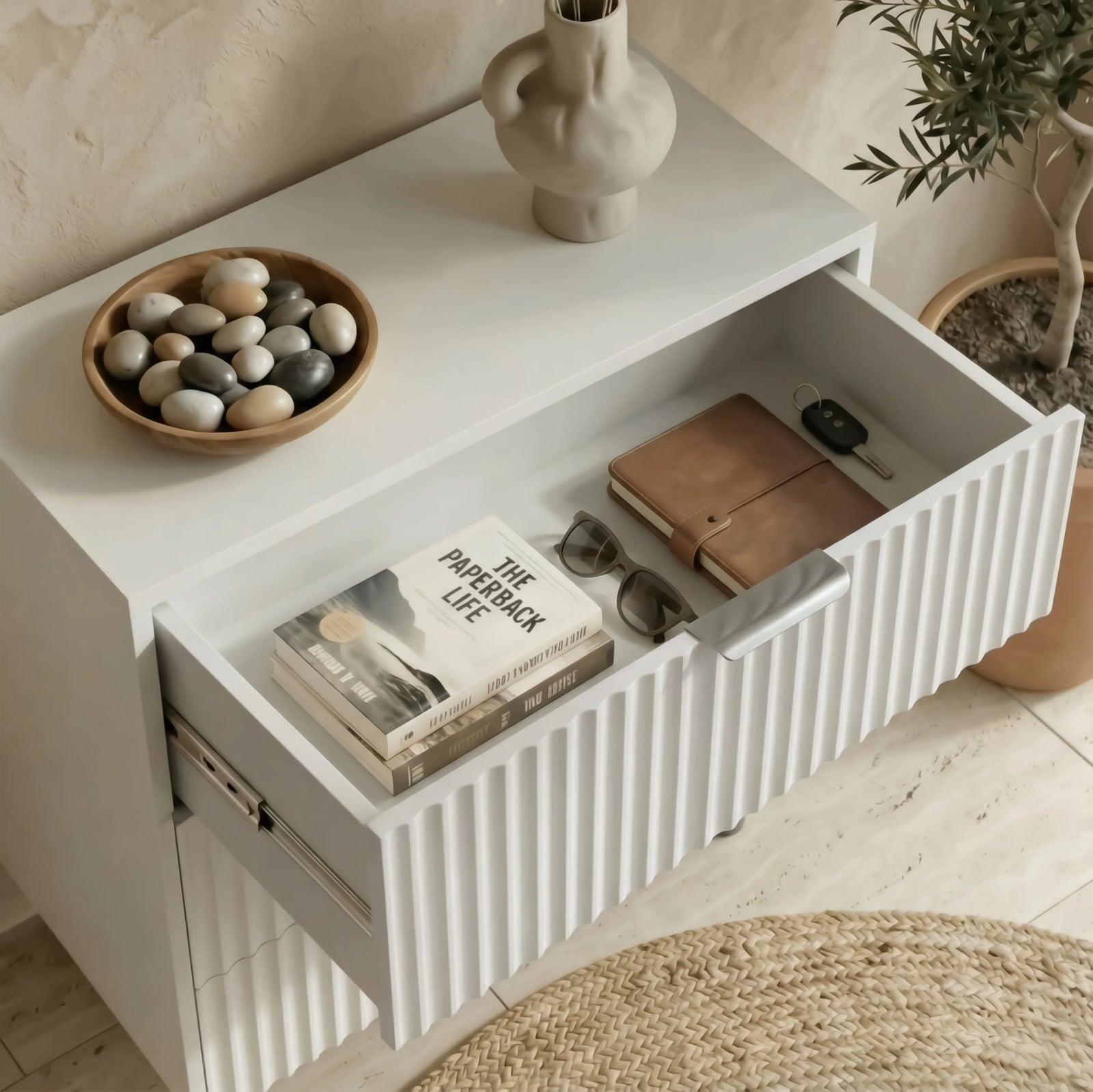 [White] Minimalist white bedside table with an open drawer containing neatly arranged books and small items