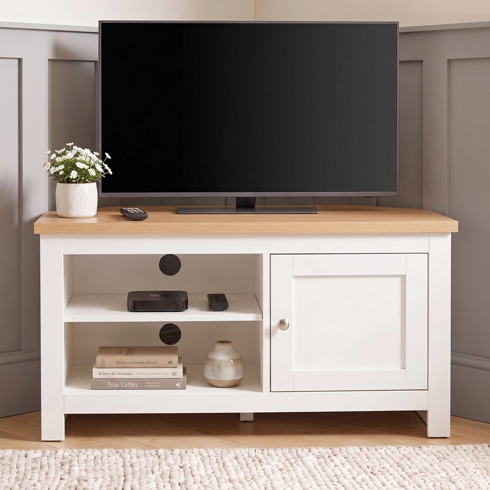 [White] Corner TV stand with a light wood top, styled with a flat-screen television, a small potted plant, and decorative items, set against a grey panelled wall and light wood floor.