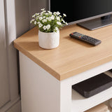 [White] close-up of a light wood corner TV stand or console table with a white cabinet base, styled with a small white flower pot and a TV remote on top.
