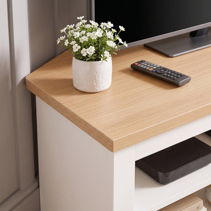 [White] close-up of a light wood corner TV stand or console table with a white cabinet base, styled with a small white flower pot and a TV remote on top.
