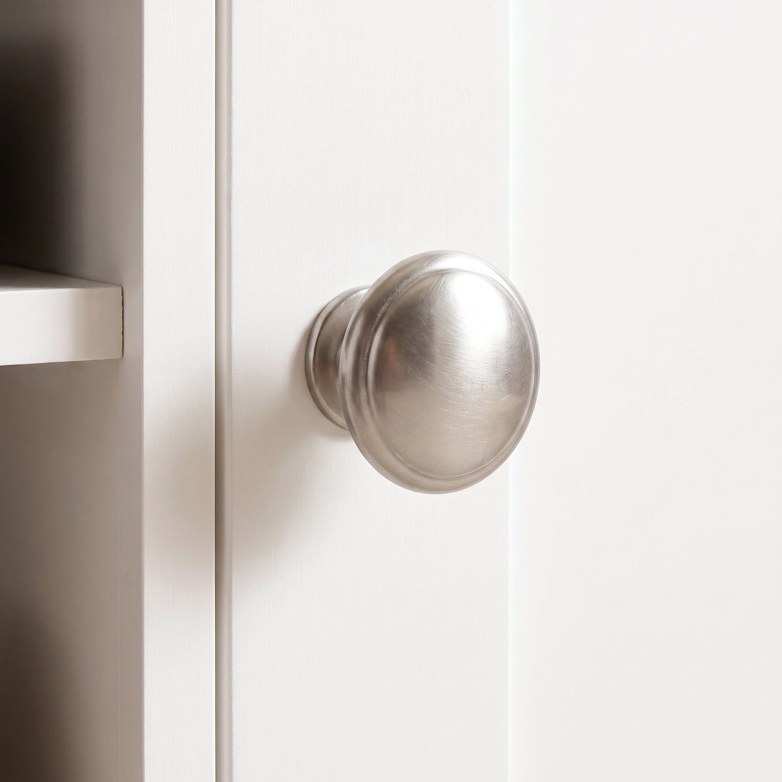 [White] round brushed metal cabinet knob mounted on a white paneled door of corner TV unit.
