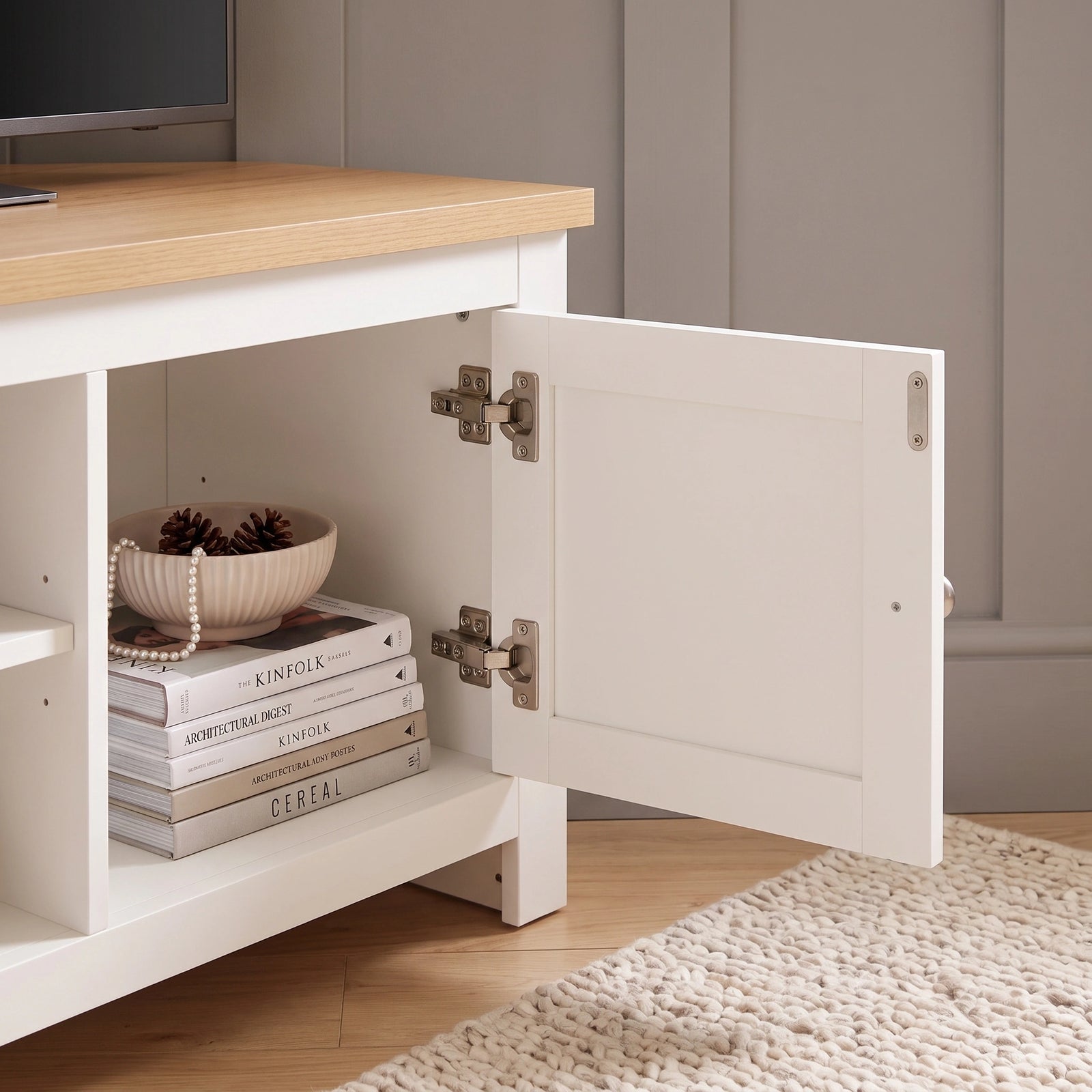 [White] Corner Tv unit console with a light oak top, shown with one cabinet door open to reveal storage space with books and a decorative bowl. The console is placed in a room setting with a light wood floor and a cream rug.