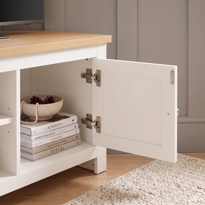 [White] Corner Tv unit console with a light oak top, shown with one cabinet door open to reveal storage space with books and a decorative bowl. The console is placed in a room setting with a light wood floor and a cream rug.