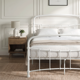 [White | Double] Close view of the white curved front rail, vertical bars and white bedding beside a small table.