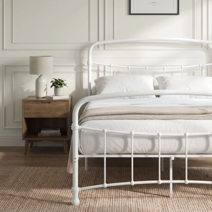 [White | Double] Close view of the white curved front rail, vertical bars and white bedding beside a small table.