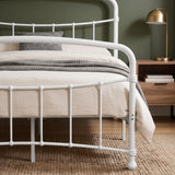[White | Double] Footboard close-up showing curved rail, bars, rounded joints, bolts, bedding, knitted throw, and lamp.