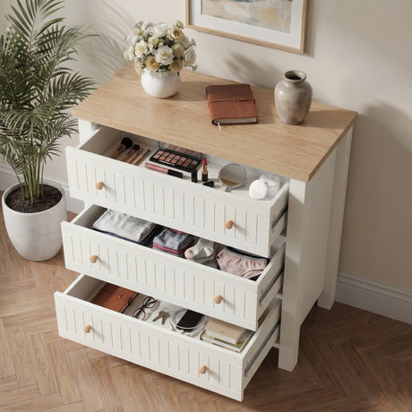 [White] dresser with open drawers containing various items, on a wooden floor with a plant and framed picture in the background