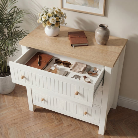[White] dresser chest with open drawer displaying items, on a wooden floor with a plant and framed picture in the background