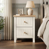[White] Linra 2 drawer bedside table beside an upholstered bed in warm bedroom lighting with ceramic lamp and vase on oak top