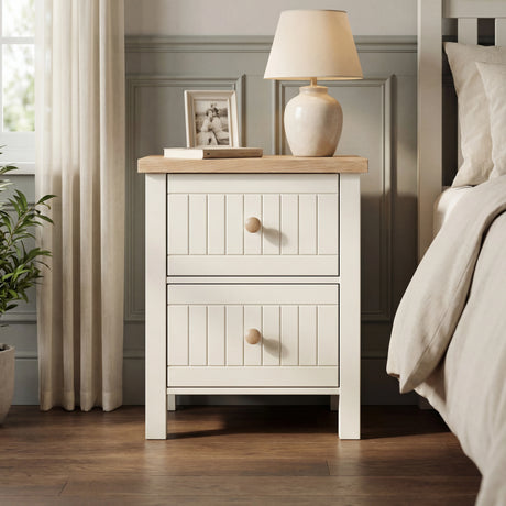 [White] Linra 2 drawer bedside table beside an upholstered bed in warm bedroom lighting with ceramic lamp and vase on oak top