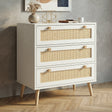 [White] Rattan three-drawer chest shown at an angle, styled with dried botanicals and gold geometric decor on top
