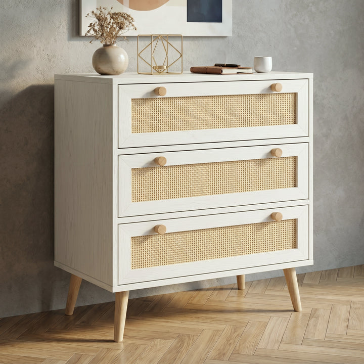 [White] Rattan three-drawer chest shown at an angle, styled with dried botanicals and gold geometric decor on top