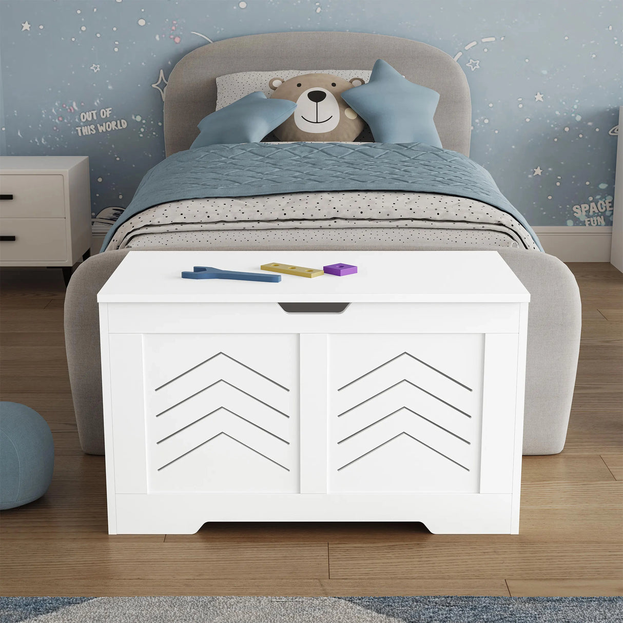 White toy storage chest in front of a child's bed with blue bedding and teddy bear.