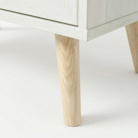 [White] Wovena close-up of a solid tapered natural wood leg showing clear grain detail where it meets the whitewashed cabinet base, with a second leg visible behind