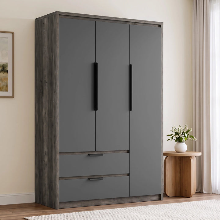 [Wooden Grey] tall wooden grey three-door wardrobe with two black vertical handles and two lower drawers, shown in a styled bedroom setting next to a wooden side table and window curtains.