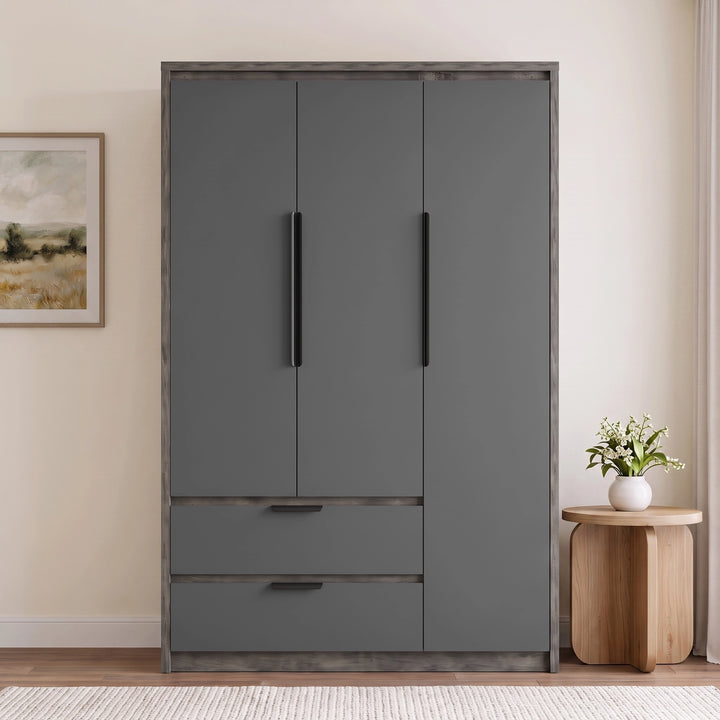 [Wooden Grey] three-door wardrobe with a matte dark gray finish and a rustic weathered wood frame, shown in a styled room setting next to a wooden side table with a vase of flowers.