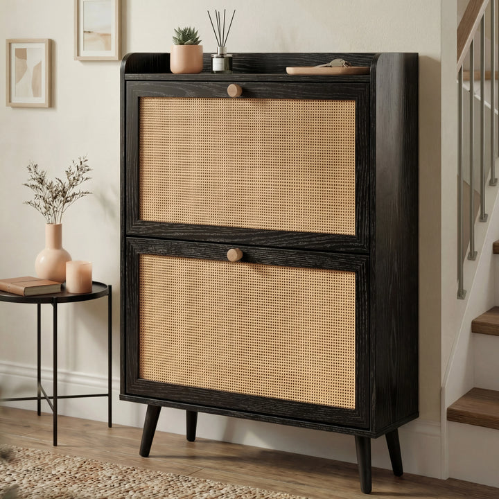 [Black] Wovena Rattan Black 2 Door Shoe Storage Cabinet angled in neutral hallway with staircase, black side table, peach vase, and succulent and reed diffuser on top shelf