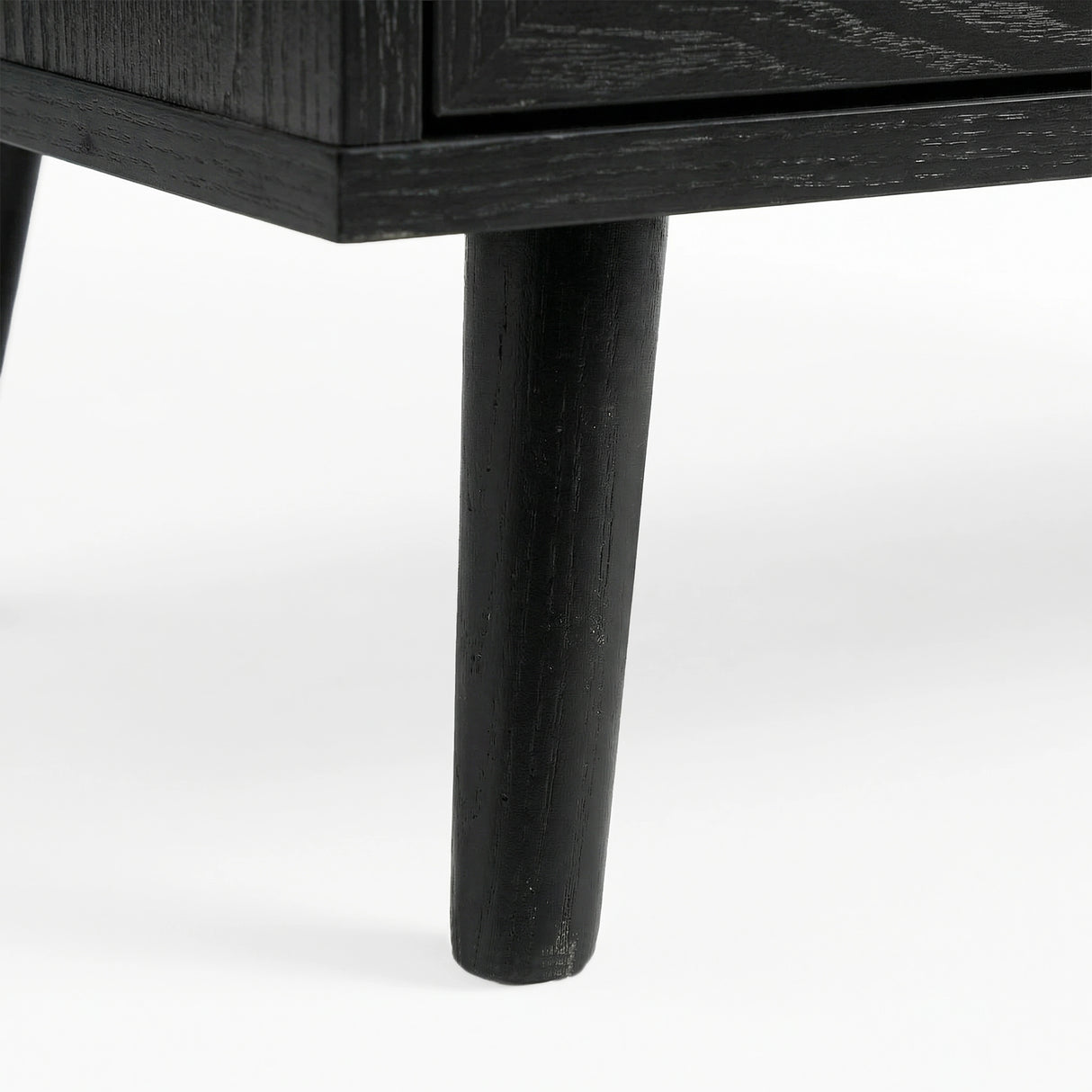 [Black] Close-up of black-painted tapered leg supporting Wovena rattan chest, wood grain visible through finish