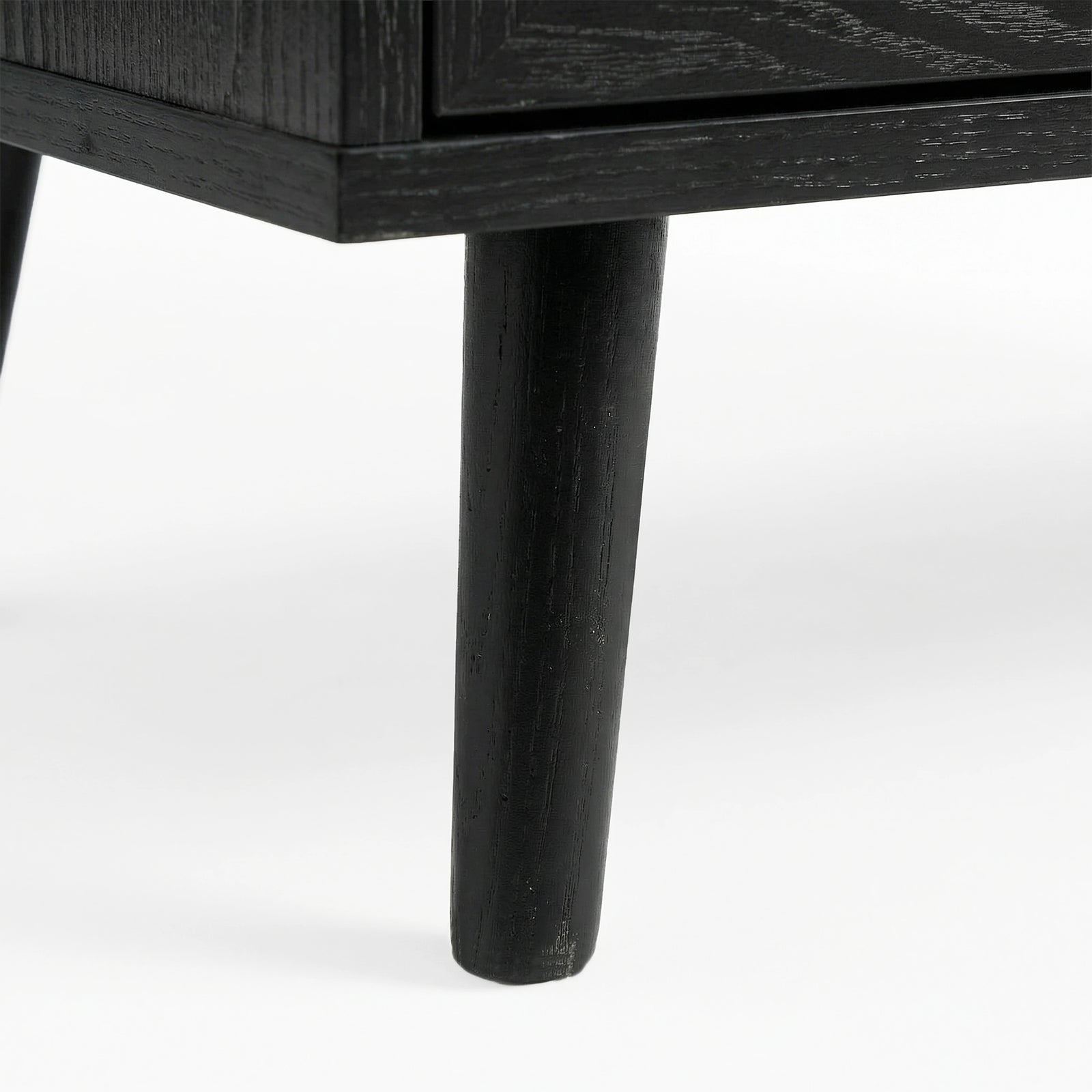 [Black] Close-up of black-painted tapered leg supporting Wovena rattan chest, wood grain visible through finish