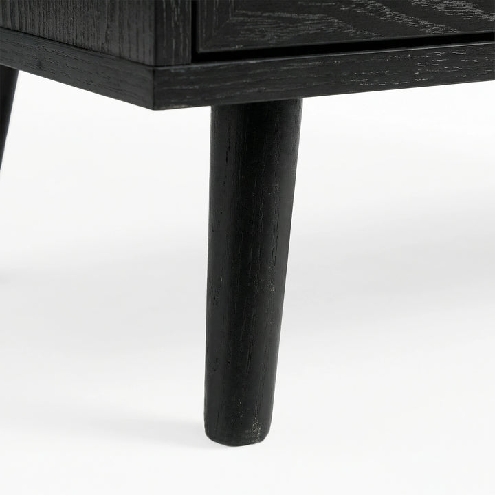 [Black] Close-up of black-painted tapered leg supporting Wovena rattan chest, wood grain visible through finish