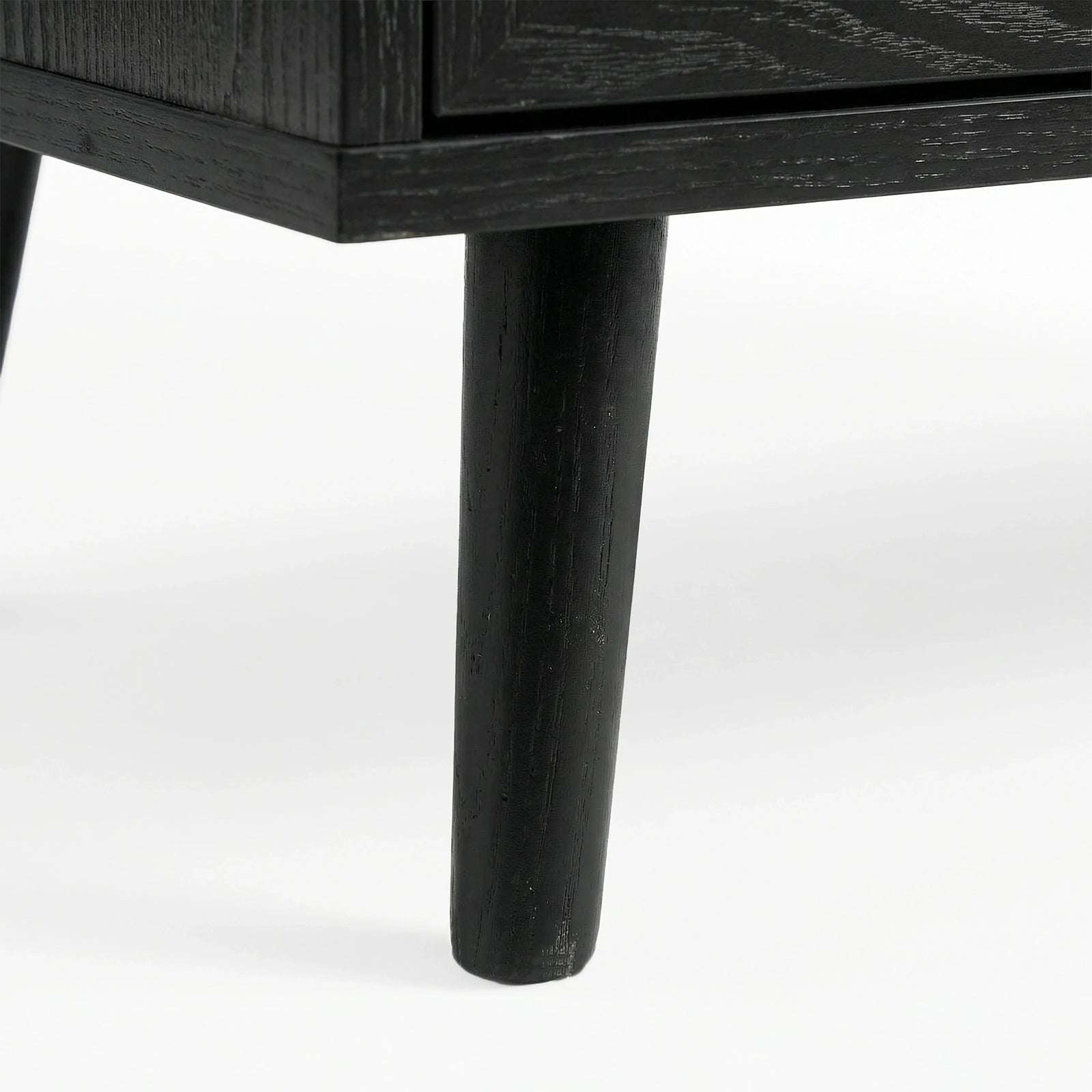 [Black] Close-up of black-painted tapered leg supporting Wovena rattan chest, wood grain visible through finish