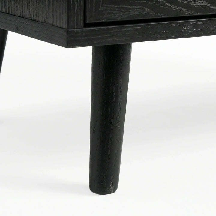 [Black] Close-up of black-painted tapered leg supporting Wovena rattan chest, wood grain visible through finish