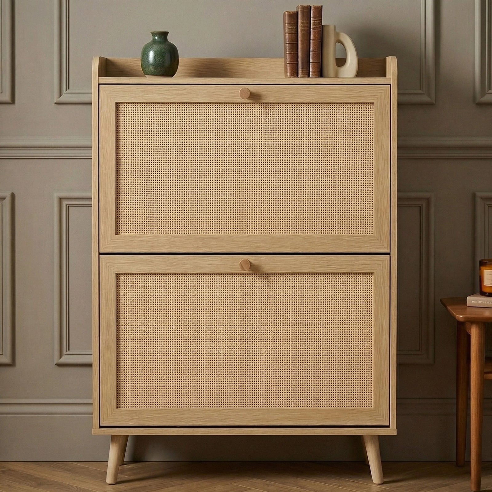 [Oak] Wovena Rattan Oak 2 Door Shoe Storage Cabinet front-facing against grey panelled wall on herringbone floor with green ceramic vase and stacked leather books on top shelf