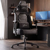 [Black] gaming chair with white XTRAKIR logos on the headrest and lumbar support, positioned in a modern home office setup with a desk, dual monitors, and wood flooring.
